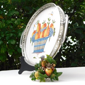 May include: A silver serving tray with a white ceramic insert featuring a hand-painted design of a blue basket filled with orange and yellow fruit. The tray has a decorative silver border and two handles.