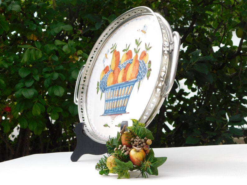 May include: A silver serving tray with a white ceramic insert featuring a hand-painted design of a blue basket filled with orange and yellow fruit. The tray has a decorative silver border and two handles.