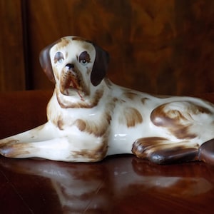 Puede incluir: Una figurita de cerámica de un perro marrón y blanco acostado. El perro tiene orejas marrones y una nariz marrón.