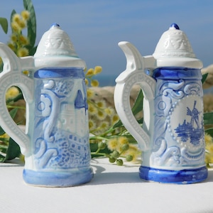 Puede incluir: Dos jarras de cerveza de cerámica azul y blanca con diseños intrincados. Una jarra presenta un molino de viento y la otra tiene un patrón floral.