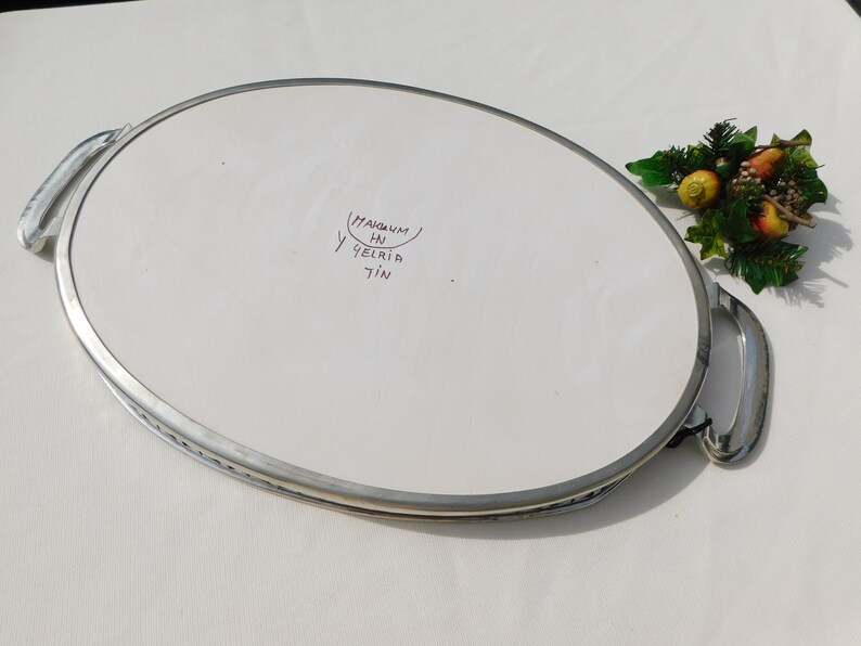 May include: A silver oval serving tray with two handles. The tray has a white ceramic insert with the words "Magnum HN 9elria 7in" written in purple ink.