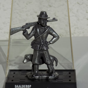 May include: A pewter figurine of a man in a hat holding a rifle and a dog. The figurine is displayed in a clear plastic case with a black base that reads "DAALDEROP Royal Holland Pewter".
