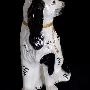 K&ouml;nnte beinhalten: Eine wei&szlig;e und schwarze Keramik-Hundefigur mit einem goldenen Halsband und einer Kette. Der Hund sitzt mit dem Kopf zur Seite gedreht und die Ohren h&auml;ngen herunter.