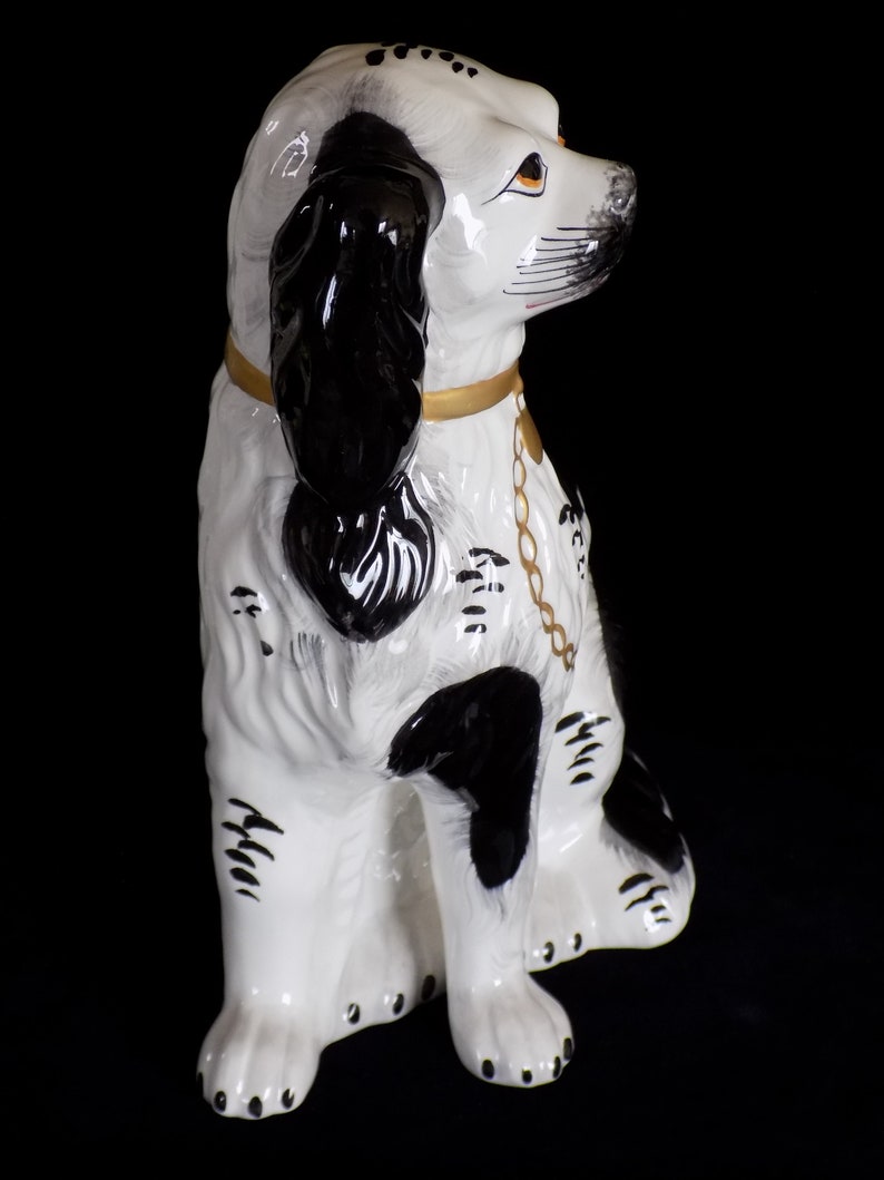 K&ouml;nnte beinhalten: Eine wei&szlig;e und schwarze Keramik-Hundefigur mit einem goldenen Halsband und einer Kette. Der Hund sitzt mit dem Kopf zur Seite gedreht und die Ohren h&auml;ngen herunter.