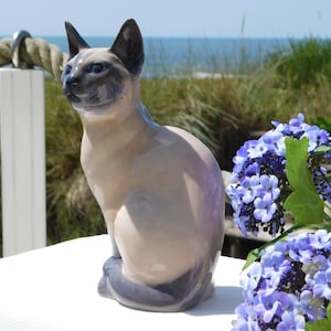 May include: A ceramic Siamese cat figurine with a glossy finish. The cat has a light tan body, dark brown ears and face, and blue eyes. It is sitting on a white surface with blue flowers in the background.