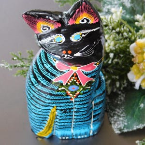 Puede incluir: Una figurita de gato de cerámica pintada a mano, negra y azul, con un lazo rosa y detalles florales coloridos. El gato tiene ojos negros con pupilas azules y una raya blanca en la nariz.