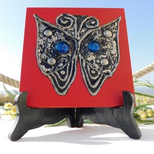 Op de afbeelding: Een metalen vlindersculptuur met blauwe glazen ogen, gemonteerd op een rood houten vierkant, tentoongesteld op een zwarte ezel.