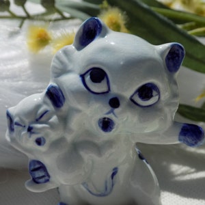 Puede incluir: Una figurita de cerámica blanca de dos gatos, uno más grande que el otro. El gato más grande está sosteniendo al gato más pequeño en sus brazos. Ambos gatos tienen ojos azules y detalles pintados en azul.