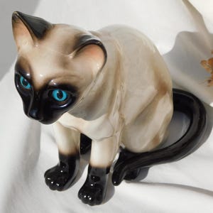 May include: A ceramic figurine of a Siamese cat with blue eyes and a black tail. The cat is sitting with its legs tucked under its body.