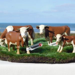 Puede incluir: Un conjunto de cuatro figuras de vacas marrones y blancas, que incluye tres vacas adultas y un ternero, sobre un trozo de hierba artificial verde. Cada figura tiene una etiqueta blanca con el logotipo de Schleich en rojo y blanco.