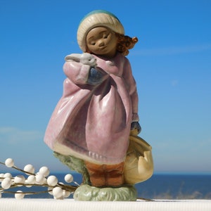 May include: A porcelain figurine of a girl wearing a pink coat and a blue and white hat, holding a white rabbit. She is standing on a green base with a brown basket.
