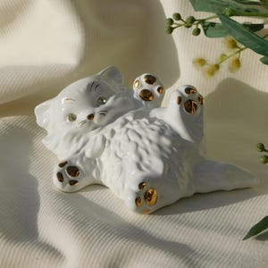 Op de afbeelding: Een witte keramische kattenfiguur die op haar rug ligt, met gouden accenten op de poten en oren. De ogen van de kat zijn versierd met kleine strass-steentjes. De figuur ligt op een witte, getextureerde achtergrond.