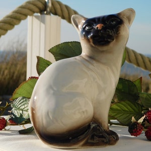 May include: A ceramic figurine of a Siamese cat sitting with its tail curled around its legs. The cat is white with black markings on its face and tail. The figurine is sitting on a white surface with green leaves and red berries.