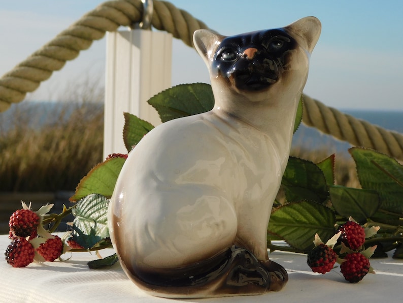 May include: A ceramic figurine of a Siamese cat sitting with its tail curled around its legs. The cat is white with black markings on its face and tail. The figurine is sitting on a white surface with green leaves and red berries.