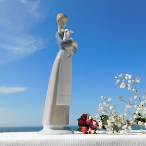 Puede incluir: Una figura de porcelana de una silueta alta y esbelta que viste una parte superior azul y una falda larga de color gris claro con delantal, sosteniendo un pequeño cordero blanco en sus brazos. La figura tiene un acabado mate y liso.