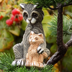 Puede incluir: Una figurita de cerámica gris y marrón de un gato con una figurita de gato marrón más pequeña acurrucada en sus brazos. El gato más grande tiene marcas blancas en la cara y las patas. Las figuritas están sentadas en una rama de pino verde.