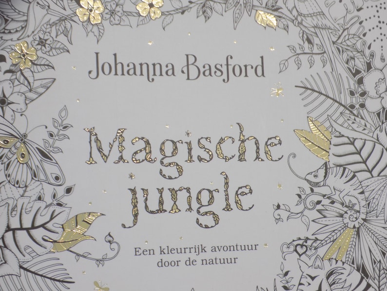 K&ouml;nnte beinhalten: Ein Buchcover mit wei&szlig;em Hintergrund und goldenen Folienakzenten. Der Titel lautet "Magische Jungle" von Johanna Basford. Der Text unter dem Titel lautet "Een kleurrijk avontuur door de natuur".