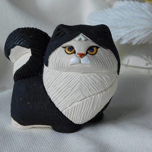 May include: A ceramic cat ornament in black and white. The cat has yellow eyes and a small orange nose. The figurine has a textured, carved appearance and a fluffy white chest. The cat's tail is curled and striped.