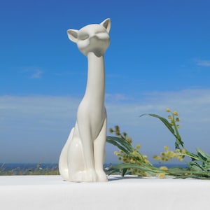 Puede incluir: Figura de cerámica blanca de un gato con cuello largo y ojos cerrados. El gato está sentado con las patas metidas debajo del cuerpo. La figura está sobre una superficie blanca con plantas verdes en el fondo.