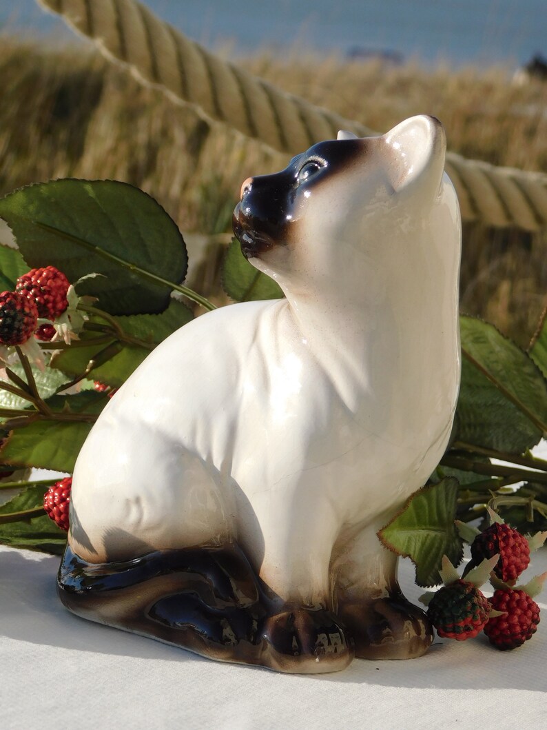 May include: A ceramic figurine of a Siamese cat sitting with its head tilted up. The cat is white with black markings on its face, ears, and tail. The figurine is painted in a realistic style.