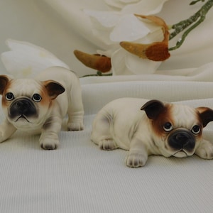 May include: Two ceramic bulldog figurines. One is standing, the other is lying down. Both have white bodies, brown ears, and black faces. The figurines are on a white surface with a floral arrangement in the background.