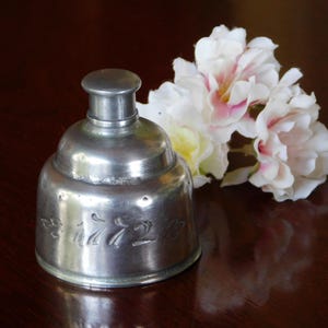 May include: A vintage, silver-toned inkwell with embossed detailing, next to a cluster of white and pink artificial flowers. The inkwell has a rounded base and a small, cylindrical lid. The flowers add a touch of color and softness to the scene.
