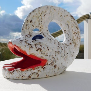 Puede incluir: Una escultura de cerámica caprichosa que se asemeja a una criatura marina estilizada. La escultura es principalmente blanca con motas marrones, una boca roja y un ojo azul. La boca abierta revela un interior rojo.