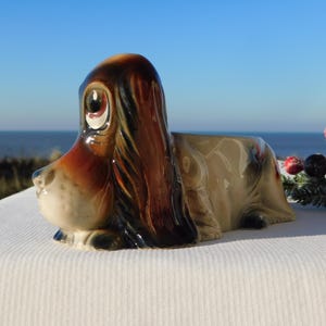 May include: A ceramic dog planter in a lying down position. The dog has brown and tan fur with large, expressive eyes. The planter is set against a backdrop of the ocean and sky, with decorative berries.