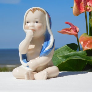 May include: A porcelain figurine of a baby wearing a blue and white hooded towel. The baby is sitting with its hand on its chin, looking to the side. The figurine is on a white surface with a blurred background of a beach and flowers.