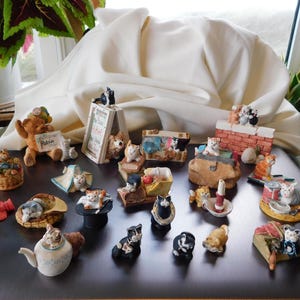Puede incluir: Una colección de figuritas de gatos en varias poses y escenarios. Las figuritas son de cerámica y presentan una variedad de colores, incluyendo blanco, naranja, negro y gris. Algunos gatos están en cestas, sobre almohadas o encima de estructuras en miniatura.