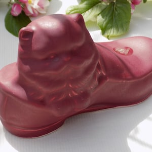 Könnte beinhalten: Ein burgunderfarbenes Keramik-Sparschwein in Form eines Schuhs. Das Sparschwein hat eine kleine Öffnung oben für Münzen.