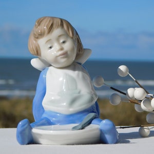 Op de afbeelding: Een porseleinen beeldje van een kind dat een blauwe slabbetje draagt en op een wit bordje zit met een blauwe lepel. Het beeldje zit met gekruiste benen. Het kind heeft bruin haar en kijkt naar de lepel.