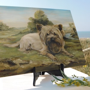 May include: A painting of a small, light brown dog with a fluffy coat, lying down in a grassy field. The painting is on a wooden easel and is set up outdoors.