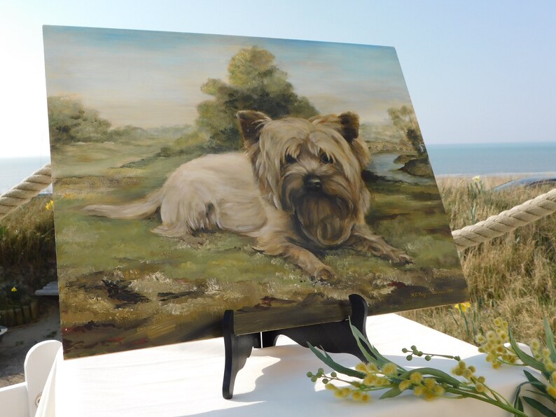 May include: A painting of a small, light brown dog with a fluffy coat, lying down in a grassy field. The painting is on a wooden easel and is set up outdoors.