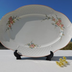 May include: White oval serving platter with a raised woven pattern and pink and blue floral design. The platter has a pink rim and is displayed on two black stands.