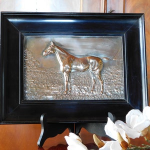 May include: A silver-toned framed relief sculpture of a horse standing in a field. The frame is black and the horse is depicted in a realistic style.