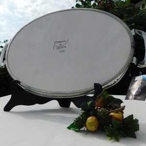 May include: A silver oval serving tray with two handles. The tray is on a black stand and has the words "MAGNUM Y DELIRIA TIN" written on the bottom.