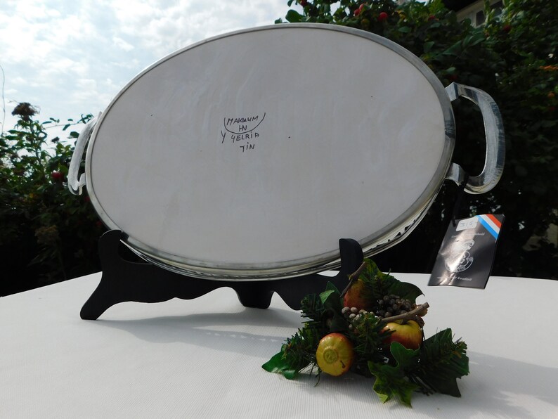 May include: A silver oval serving tray with two handles. The tray is on a black stand and has the words "MAGNUM Y DELIRIA TIN" written on the bottom.