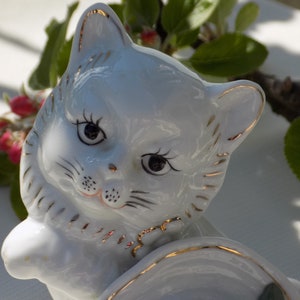 以下が含まれることがあります： 金色のアクセントが付いた白い磁器製の猫の置物。猫は頭を傾け、目を閉じている。置物はヴィンテージスタイルで、繊細な外観をしている。