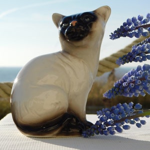 May include: A ceramic figurine of a Siamese cat sitting with its tail curled around its legs. The cat is white with black markings on its face and tail. The figurine is sitting on a white surface with blue flowers in the background.