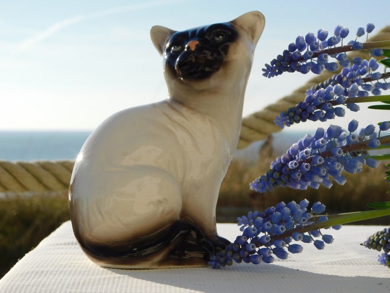 May include: A ceramic figurine of a Siamese cat sitting with its tail curled around its legs. The cat is white with black markings on its face and tail. The figurine is sitting on a white surface with blue flowers in the background.