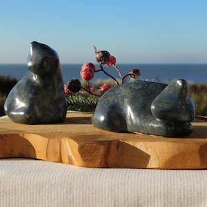 Op de afbeelding: Twee donkerblauwe stenen konijnsculpturen, één rechtop en één liggend, staan op een natuurlijke houten plank. De sculpturen hebben een glanzende afwerking en staan voor een zeegezicht met een helderblauwe lucht. Decoratieve bessen zijn op de achtergrond.