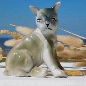 May include: A ceramic figurine of a seated dog with a slightly grumpy expression. The dog is primarily gray with white accents and a glossy finish. The background features dried grasses and a blue sky.