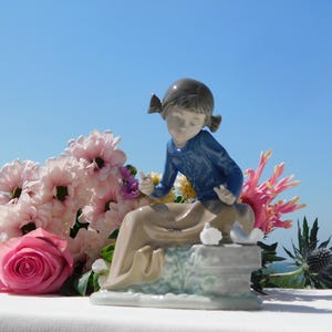 May include: A porcelain figurine of a young person with dark hair in pigtails, wearing a blue long-sleeved top and a long tan skirt. The figure sits on a stone-like base with two small white birds, positioned among pink roses and flowers.