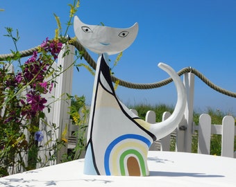 Può includere: Una scultura di gatto in ceramica bianca con un design arcobaleno sul suo corpo. Il gatto ha un contorno nero e un sorriso blu. La scultura è seduta su un tavolo bianco.