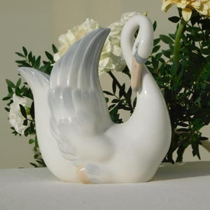 Puede incluir: Una figurita de cisne de porcelana blanca con un detalle de ala gris. El cisne está posado con la cabeza girada hacia un lado y el cuello curvado.