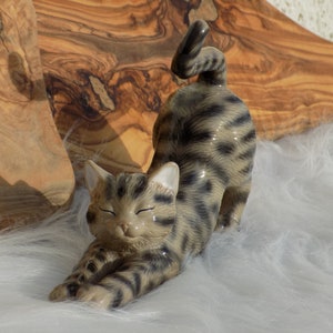 Op de afbeelding: Een keramieken beeldje van een zich uitrekkende kat met een tabby-patroon in bruin- en zwarttinten. De kat is afgebeeld met gesloten ogen, in een ontspannen houding. Het beeldje staat tegen een achtergrond van witte vacht en een houten oppervlak.