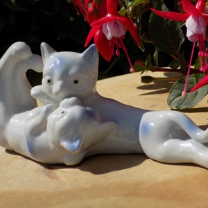 Könnte beinhalten: Eine weiße Keramikfigur von zwei Katzen, die sich kuscheln. Die Katzen sind mit einem hellblauen Glasur bemalt und haben schwarze Augen. Die Figur steht auf einer Holzoberfläche.