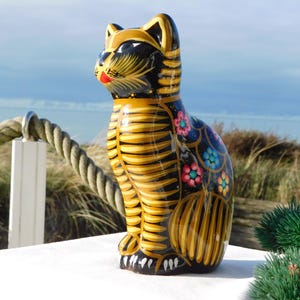 Può includere: Statuina decorativa a forma di gatto in ceramica con corpo a strisce nere e gialle, decorazioni floreali e bocca rossa. Il gatto è in posizione seduta, con la testa girata di lato. La statuina è esposta all'aperto.