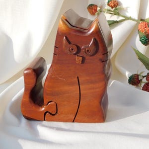 Könnte beinhalten: Handgeschnitzte Holzkatzenfigur in warmem, rotbraunem Farbton. Die Katze ist mit detaillierten Merkmalen wie großen Augen, einer kleinen Nase und einem gebogenen Schwanz geschnitzt. Ein dekorativer Artikel.